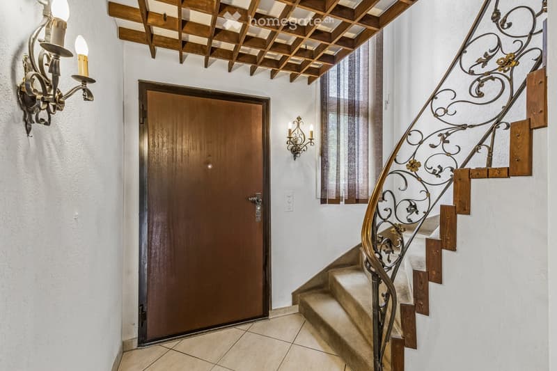 Elegant hallway with a wooden door, ornate wall-mounted candle holders, and a staircase with a decorative wrought-iron railing and wooden steps.