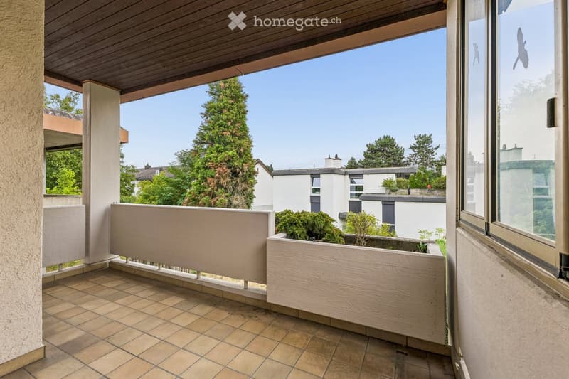 Balcony with tiled floor, wooden planter, and wooden railing, overlooking a green area with trees and neighboring houses.