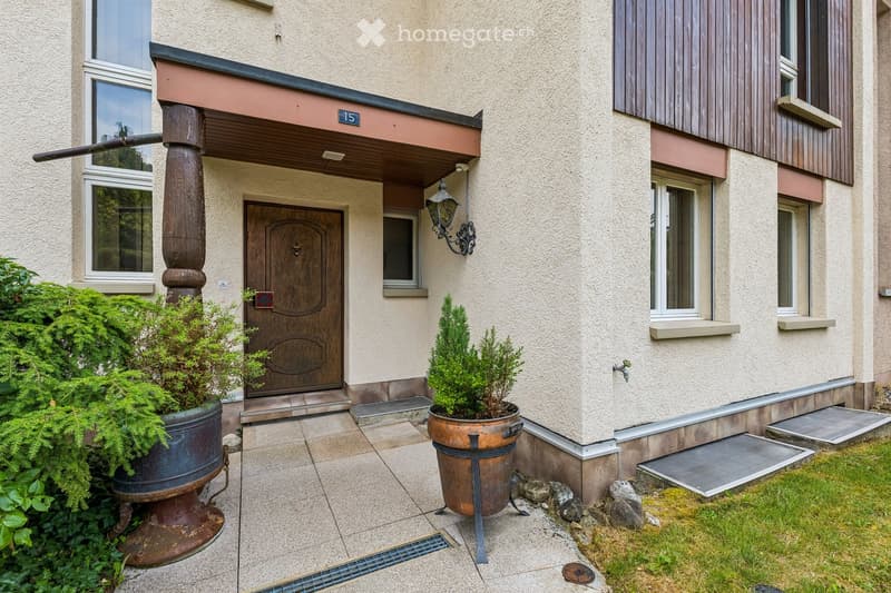 Front entrance of a two-story building with a wooden door, decorative columns, potted plants, and a small garden area.
