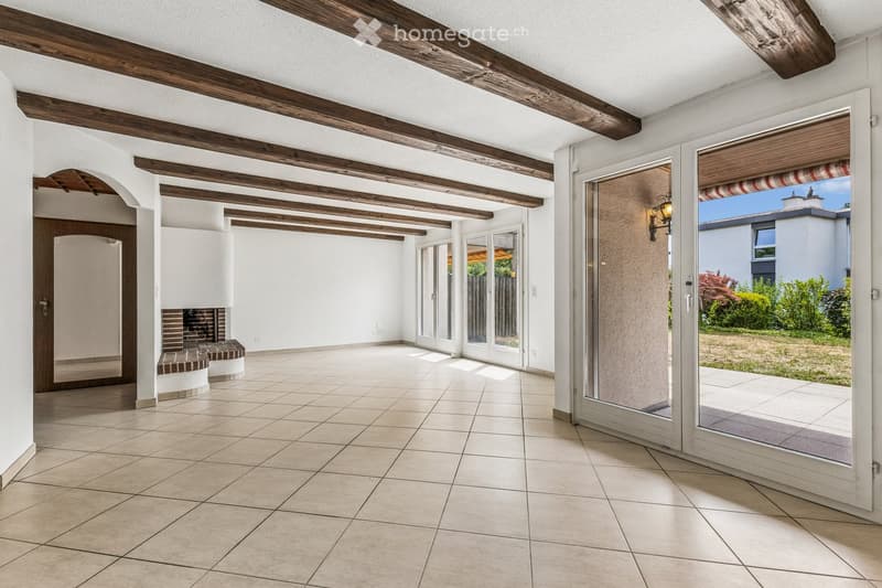 Bright, open space with exposed wooden beams, tiled floor, and large glass doors leading to an outdoor area. Features a built-in brick fireplace and white walls.