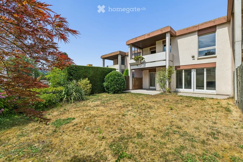 A two-story house with a modern design, featuring large windows, a balcony, and a well-maintained garden with shrubs and trees.
