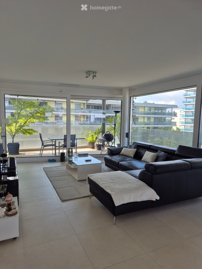 Modern living room with large windows and balcony access, featuring a black leather sofa, coffee table, ottoman, side table, and potted plants.