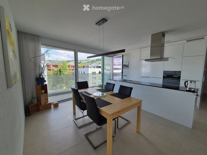 Modern open-plan kitchen and dining area with a wooden dining table, black chairs, white kitchen cabinets, and a large window overlooking a green landscape.