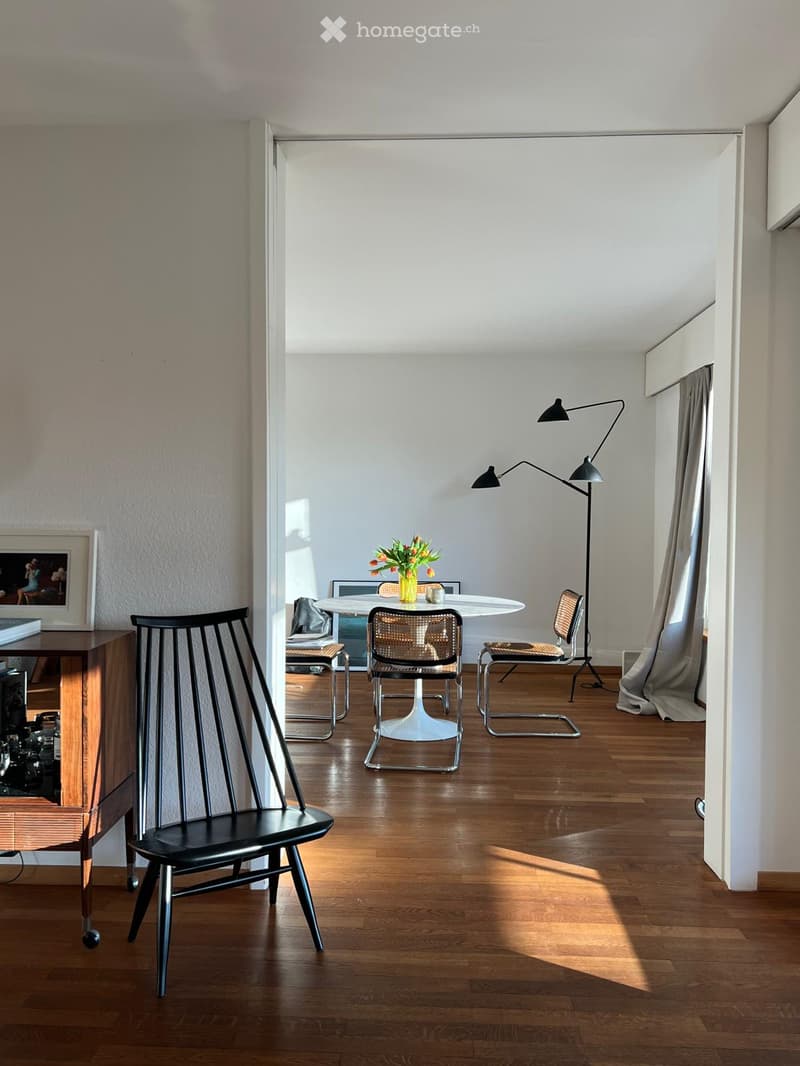 Bright, modern living space with wooden flooring, white walls, and large windows. Features a black chair, white dining table with chairs, a floor lamp, a wooden cabinet with a picture frame, and a vase with flowers on the table.