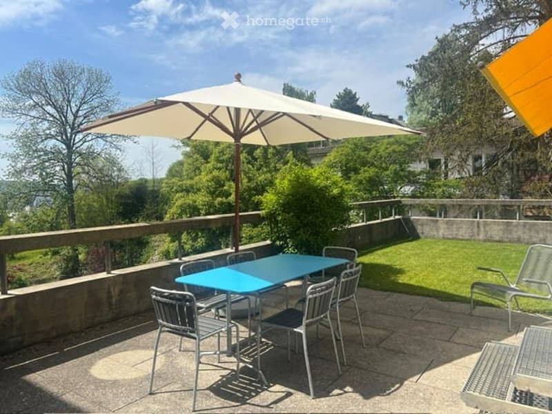 Balcony with a large umbrella, blue table, and chairs, overlooking a green lawn and trees.