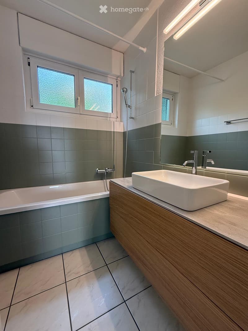 Modern bathroom with a white bathtub, a wooden vanity with a white sink, and a large mirror above the sink. The walls are tiled in grey and white, and the floor is tiled in light marble.