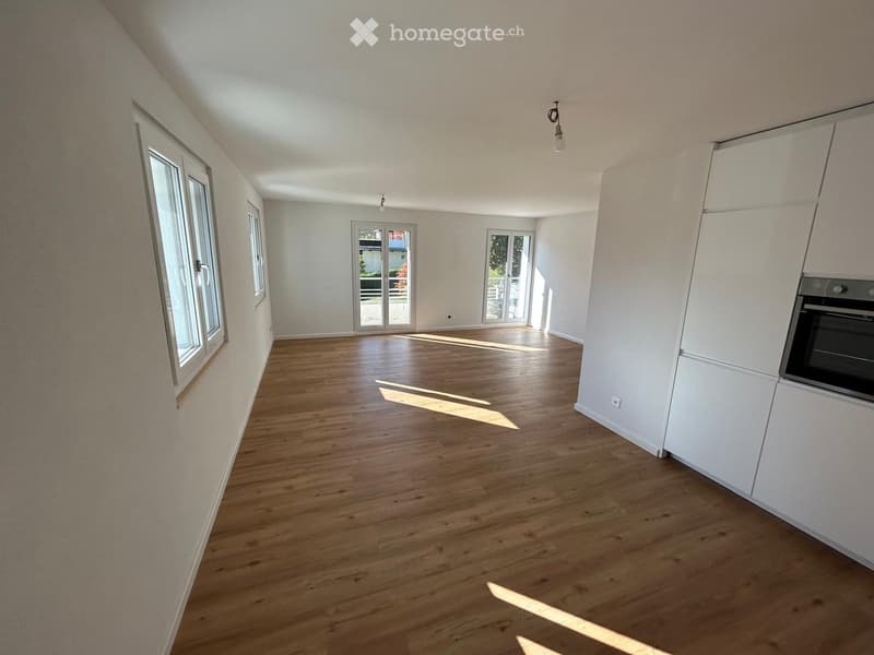 Empty room with wooden flooring, white walls, and large windows allowing natural light to enter. The room features built-in white cabinetry on one side and a partially visible oven.