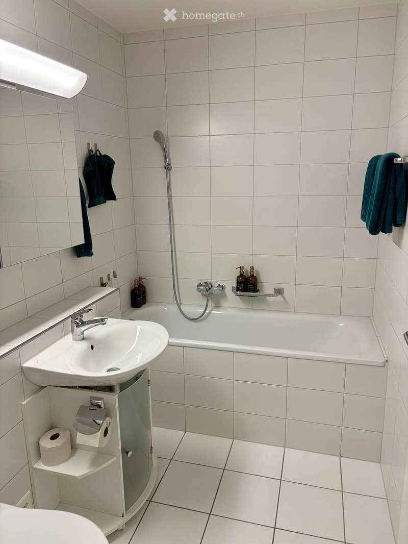 Modern bathroom with white tiles, a sink with a faucet, a bathtub with a showerhead, a mirror, and a towel rack with towels.