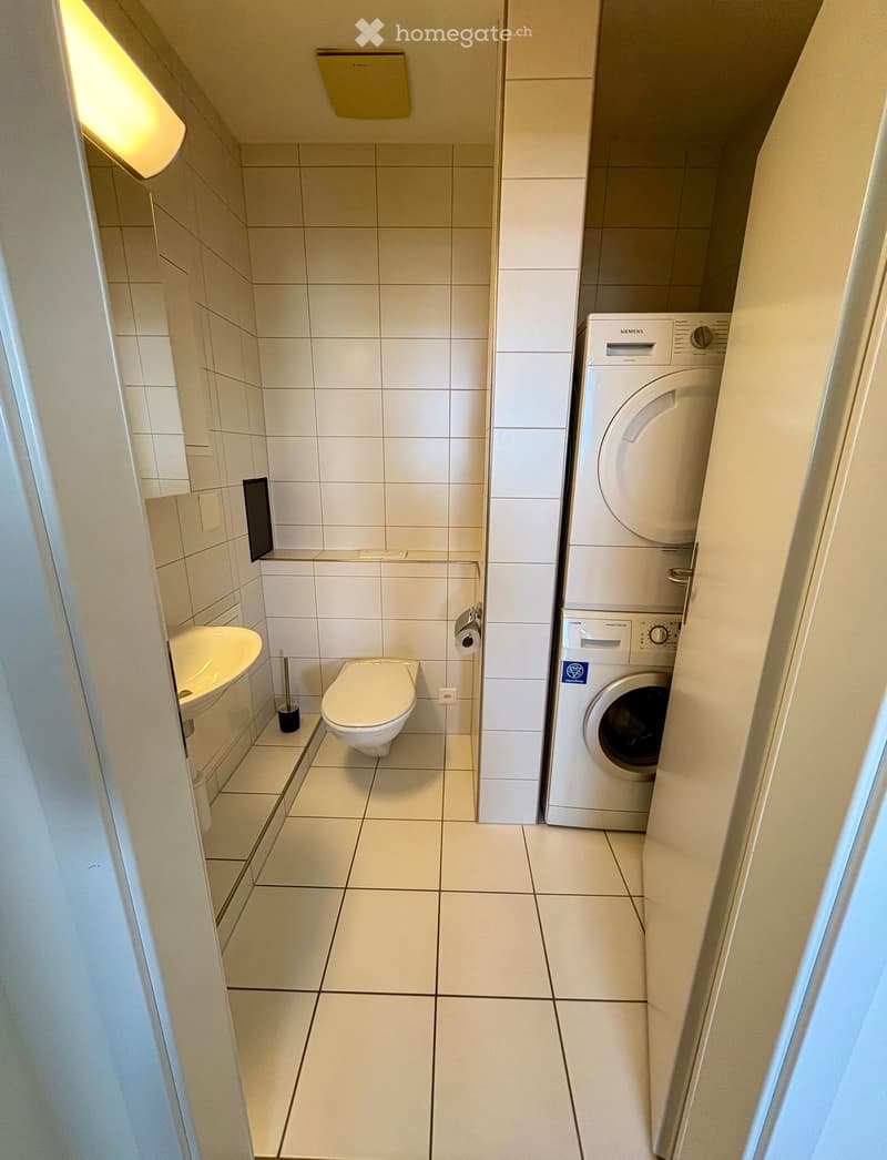 Compact bathroom with a toilet, sink, and a washing machine in the corner. The walls and floor are tiled in light beige.