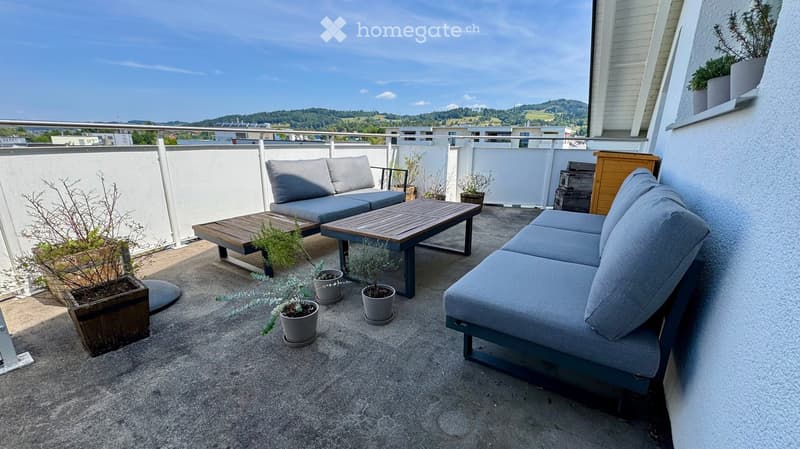 Balcony with grey sofa, wooden table, and several potted plants arranged on a concrete surface.