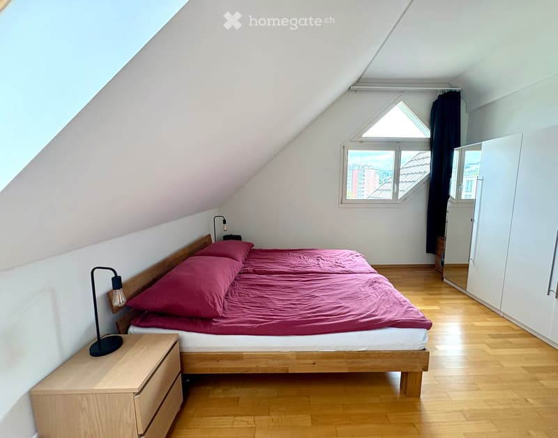Cozy attic bedroom with wooden bed, red bedding, nightstand, and lamp. Large windows offer a view of the city.