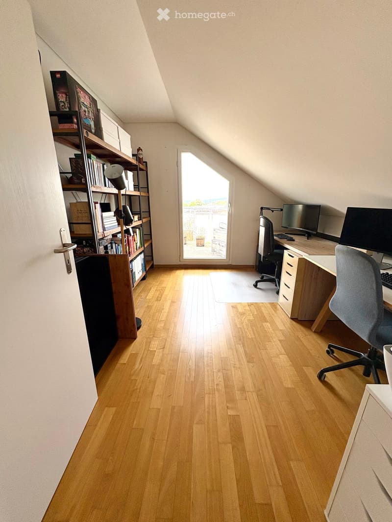 Bright attic room with wooden flooring, large window, desk, chair, and bookshelf.