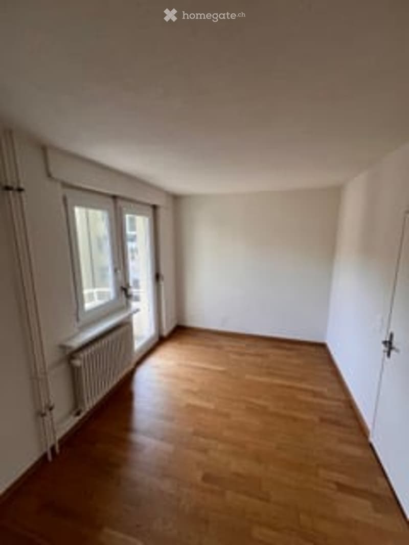 Empty room with wooden flooring, white walls, and large windows with blinds. Radiator visible under one window.
