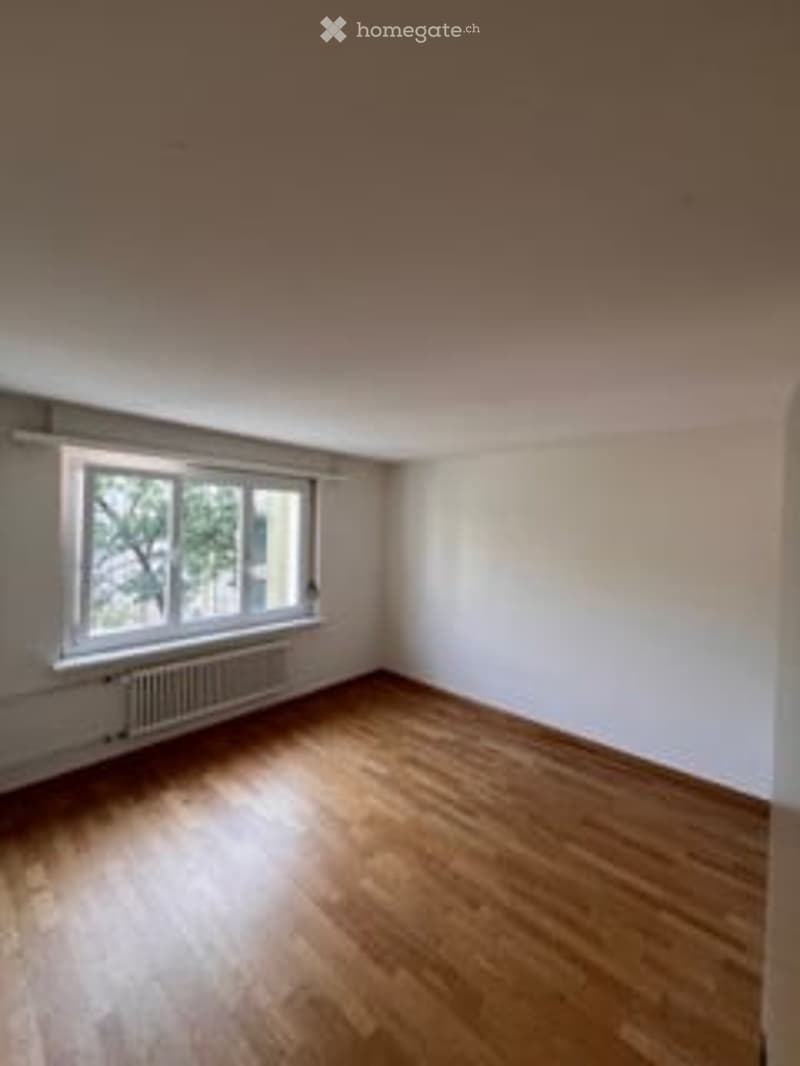 Completely empty room with wooden flooring, white walls, and large windows with radiators beneath them.
