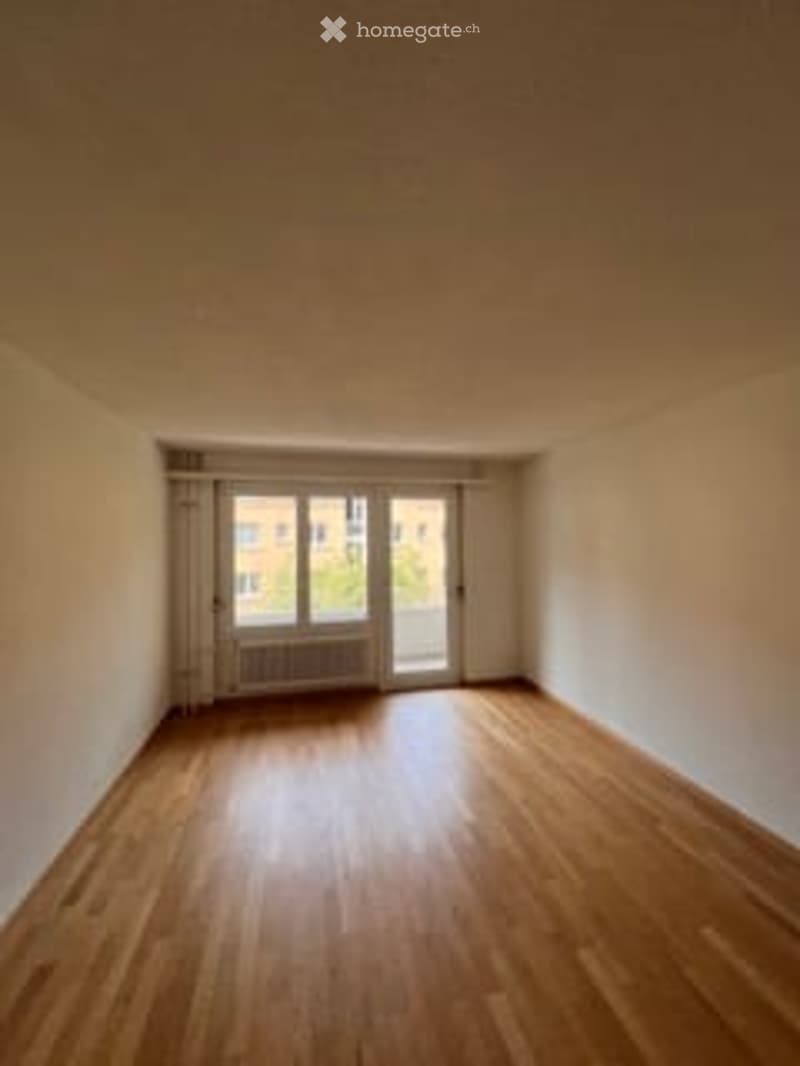 Empty room with wooden flooring, white walls, and large windows with a view of the outside.