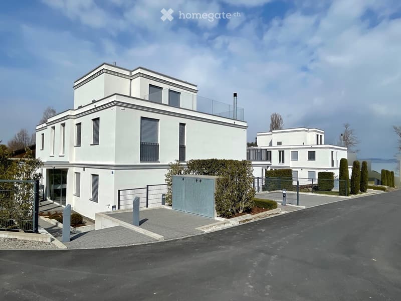 white 2 story house, with windows, glass balcony, parking place in front, paved driveway, white and gray walls, greenery