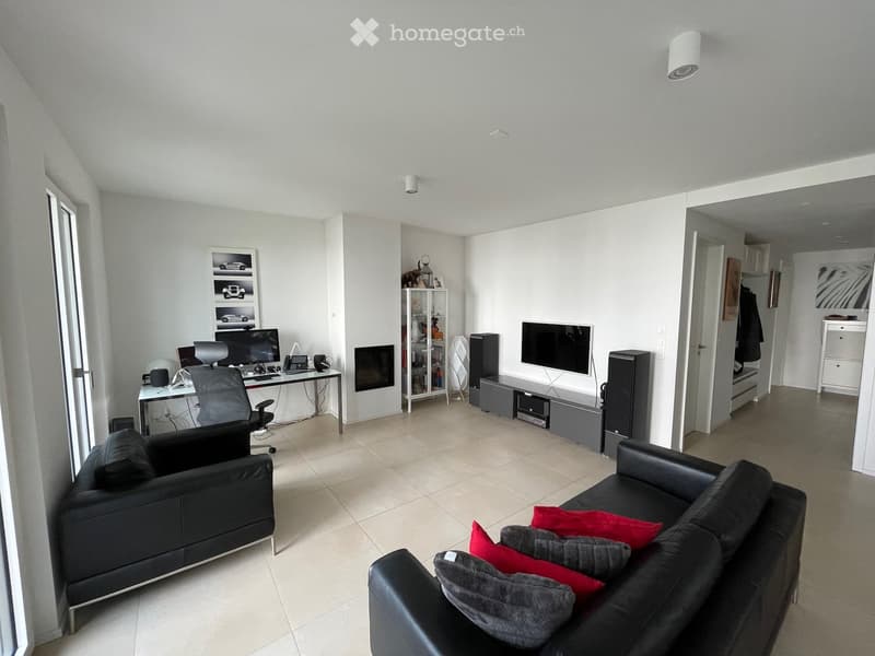 Modern living room with two black sofas, red cushions, a fireplace, a desk with a computer, and a TV on a stand.