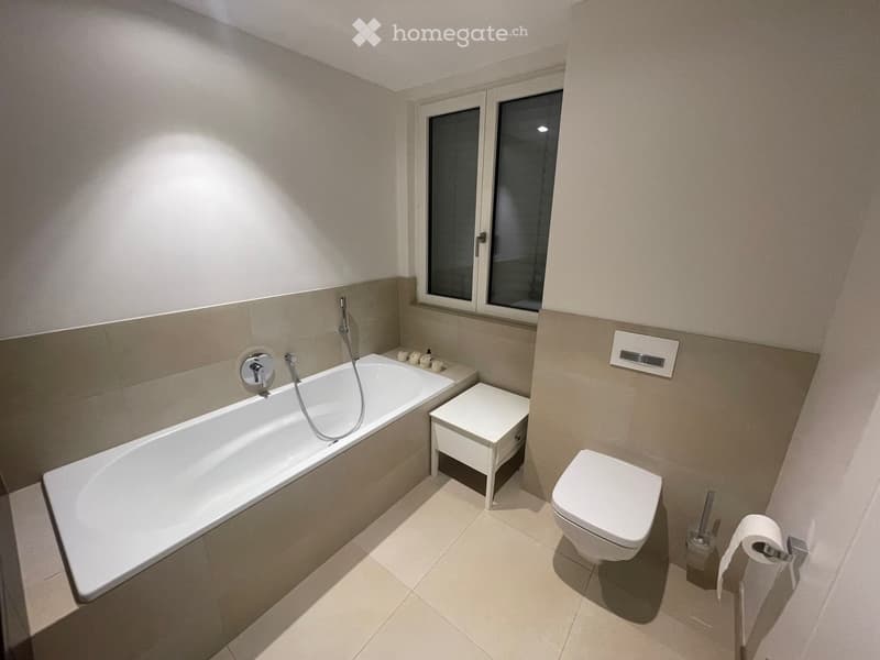 Modern bathroom with a bathtub, toilet, small stool, and toilet paper holder. Light-colored tiles and a window provide natural light.