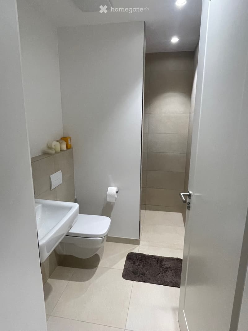 Modern bathroom with white tiles, a toilet, sink, and a small shelf holding towels and soap. A dark bathroom mat is placed on the floor near the shower area.