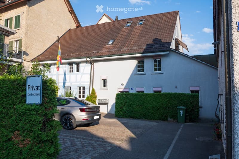 Two-story house with a brown tiled roof, white walls, multiple windows, and a flagpole with a rainbow flag. A silver car is parked in the driveway, and there is a "Privat Kein Durchgang" sign on the left side.