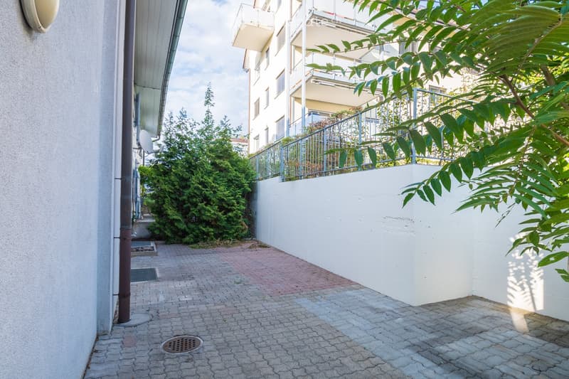A paved outdoor area with a white wall, greenery, and a metal railing leading to a balcony.