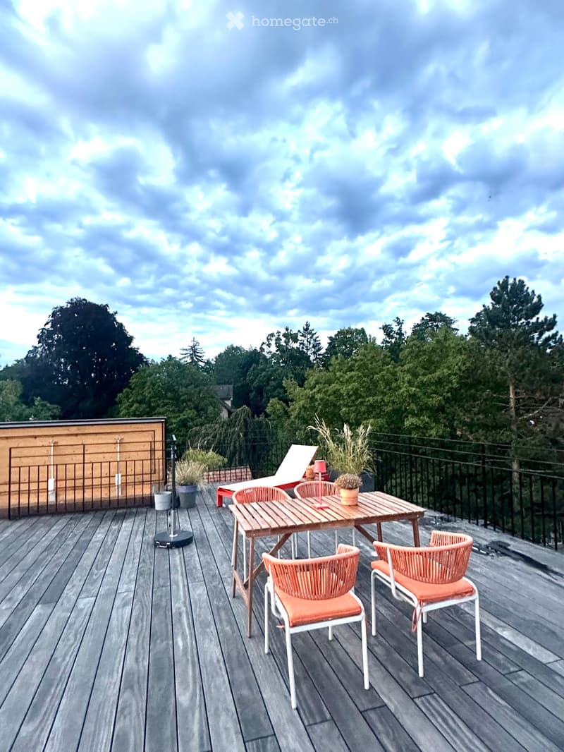 Wooden deck with a table, chairs, sun lounger, and potted plants, surrounded by a metal railing and trees.