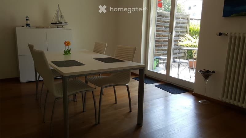 Bright dining room with a white dining table, four beige chairs, a white cabinet, a small flower vase, and a model sailboat on top of the cabinet. A large sliding glass door leads to a patio with outdoor furniture.