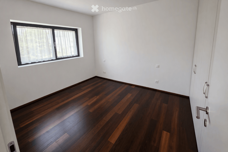 Empty room with light grey walls, dark wooden flooring, and a large window with white curtains. No furniture or equipment present.