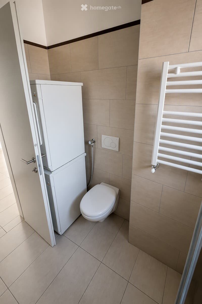 Modern bathroom with beige tiled walls and floor, white toilet, white radiator, and white cabinet.