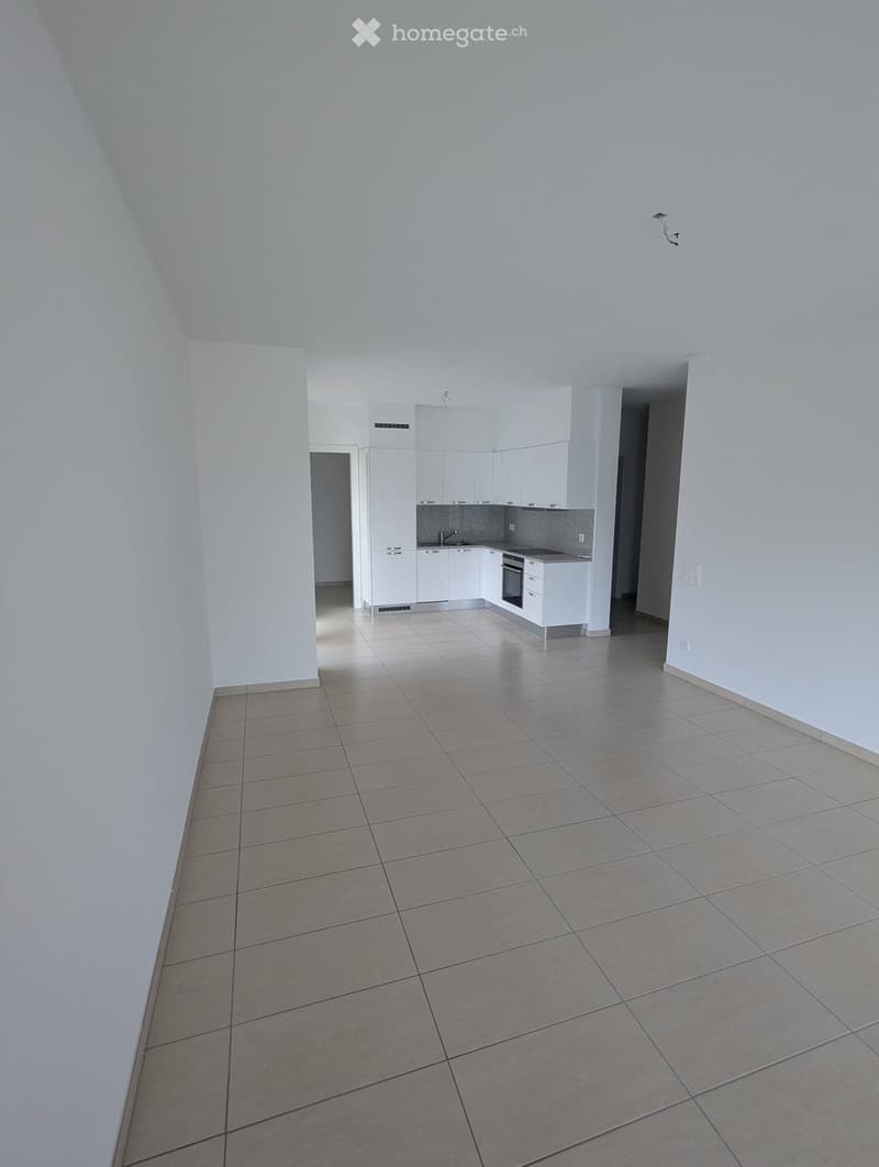 Empty room with tiled floor and white walls, featuring a small kitchen area with white cabinets and appliances.