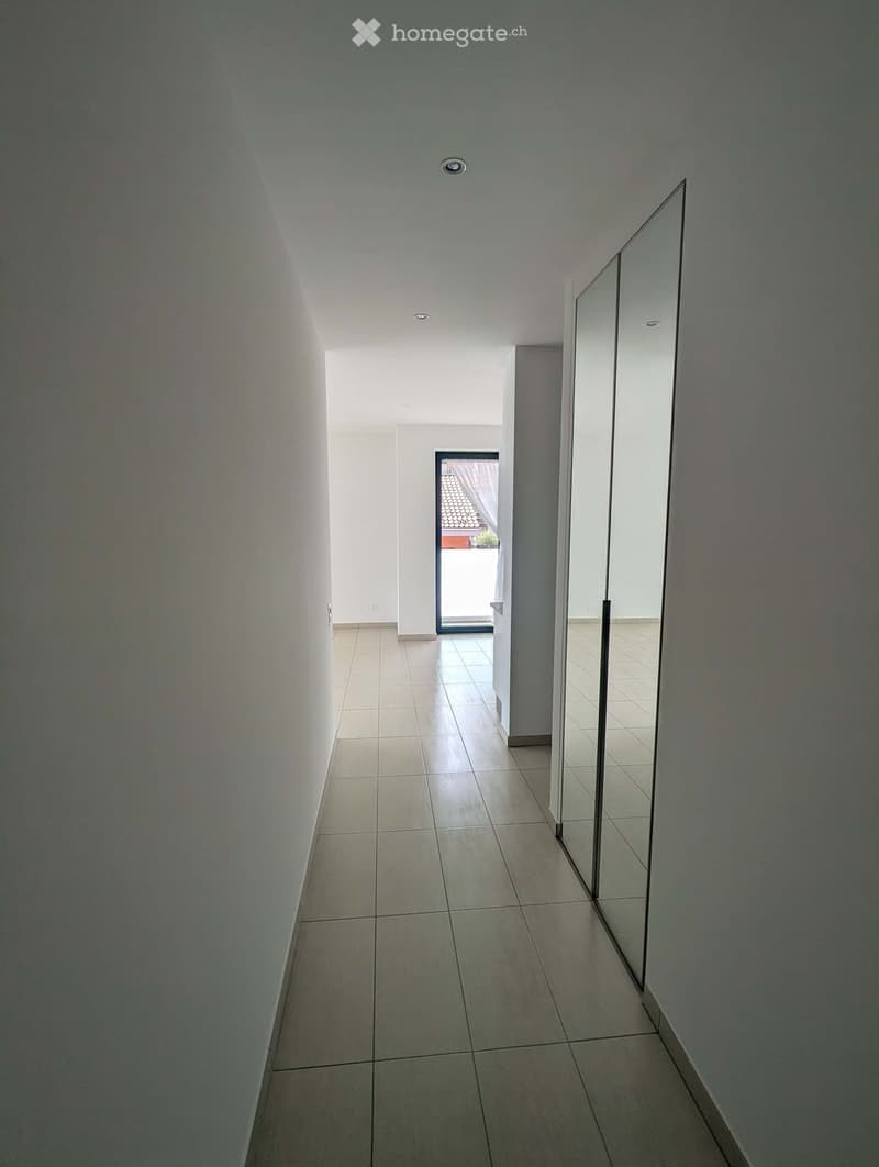 Empty hallway with tiled floor, white walls, and a large mirror on one side. Natural light enters from an open doorway at the end.