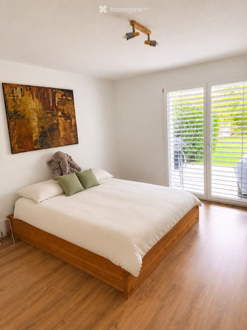 Modern bedroom with a wooden bed, white bedding, green pillows, an elephant plush toy, and a large abstract artwork on the wall. Large windows with blinds offer a view of the garden.