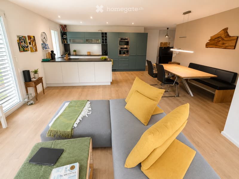 Open-plan living and dining area with a grey sofa, yellow pillows, coffee table, laptop, books, and a modern kitchen with green cabinets and stainless steel appliances.
