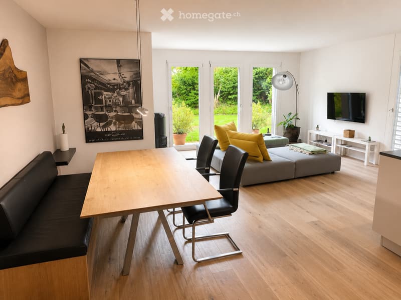 Modern living room with a wooden table, black leather bench, grey sofa with yellow cushions, black chairs, floor lamp, and a TV on the wall. Large windows provide a view of a green garden.