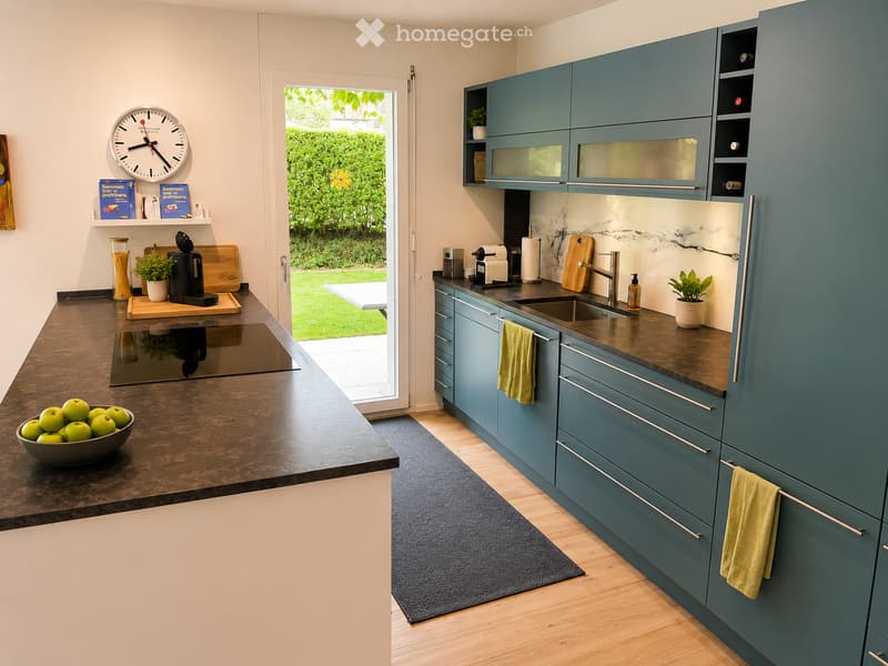 Modern kitchen with teal cabinets, dark countertops, and a large island. Features a sink, coffee maker, cutting board, bowl of apples, clock, and books on a shelf.