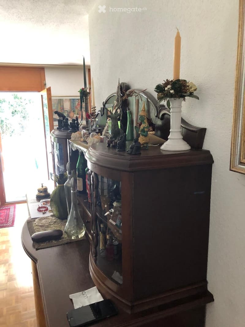 A well-lit room with a wooden cabinet, a table with decorative items including a vase, candles, and statues, and a glass cabinet with various items inside.