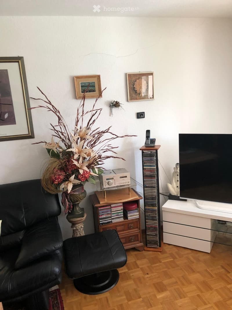 Living room with a black leather sofa, a matching leather stool, a wooden table with a flower vase, a CD rack, a TV stand with a flat-screen TV, and several picture frames on the wall.
