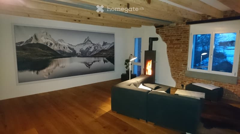 Modern living room with exposed wooden beams, large windows, a fireplace, and a large picture of a mountain landscape.