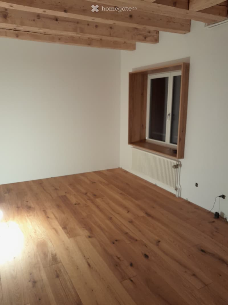 Empty room with wooden flooring, white walls, and exposed wooden beams on the ceiling. A window with a radiator below is visible on one wall.