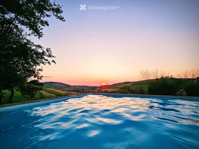 A swimming pool with a scenic view of a sunset over rolling hills and a tree-lined landscape.