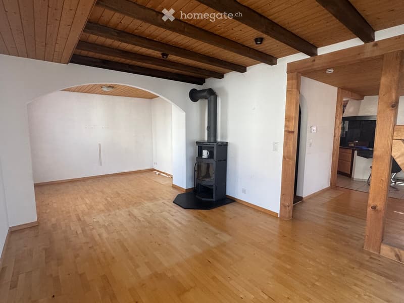 Bright living room with wooden floors, exposed wooden beams, and a black wood-burning stove. The space is open and airy with white walls and a visible kitchen area in the background.