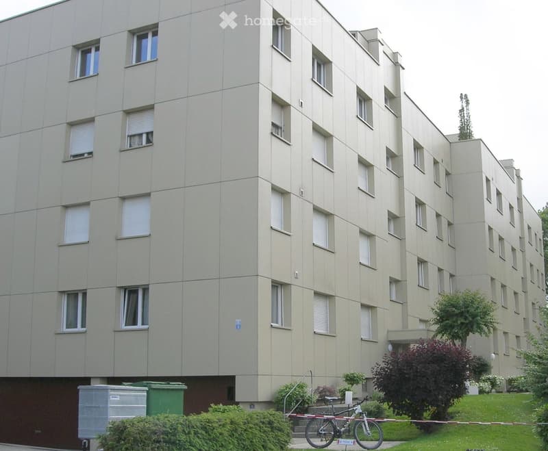 A multi-story residential building with light beige walls, numerous windows with blinds, and a small garden area with greenery and a bicycle near the entrance.