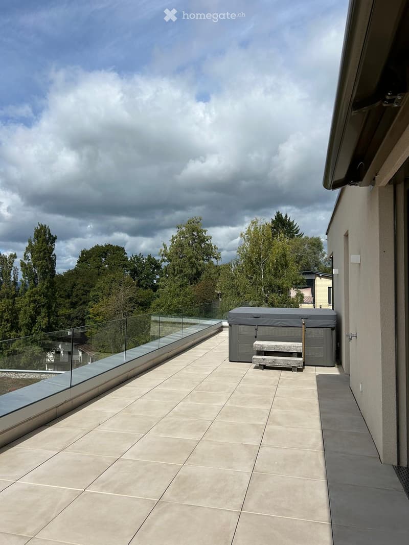 Modern balcony with tiled floor, glass railing, hot tub, and bench, overlooking a green landscape.