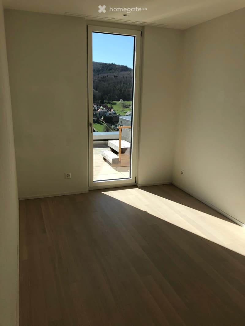 Empty room with wooden flooring, large sliding glass door leading to a balcony with a bench, and a scenic view of a green landscape.