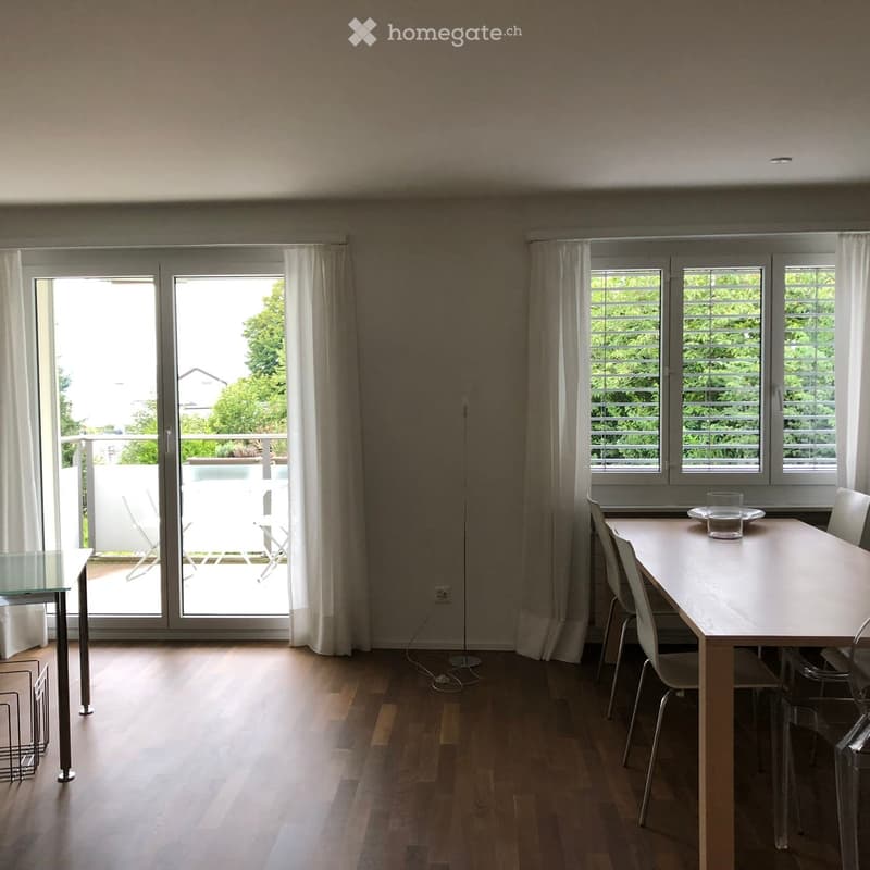 Bright dining room with wooden floor, large windows, and sliding glass doors leading to a balcony. Includes a dining table with chairs and a glass side table.