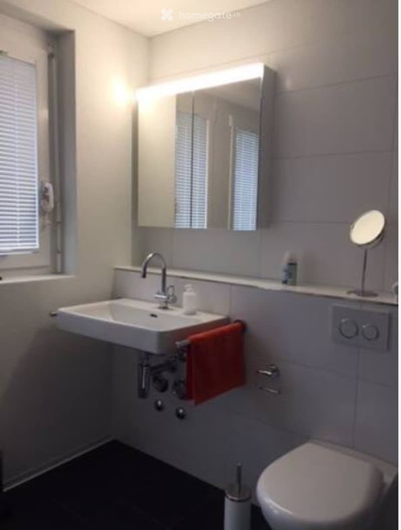 Modern bathroom with white tiles, a white sink, a chrome faucet, a large mirror with built-in lighting, a red towel, and a toilet.
