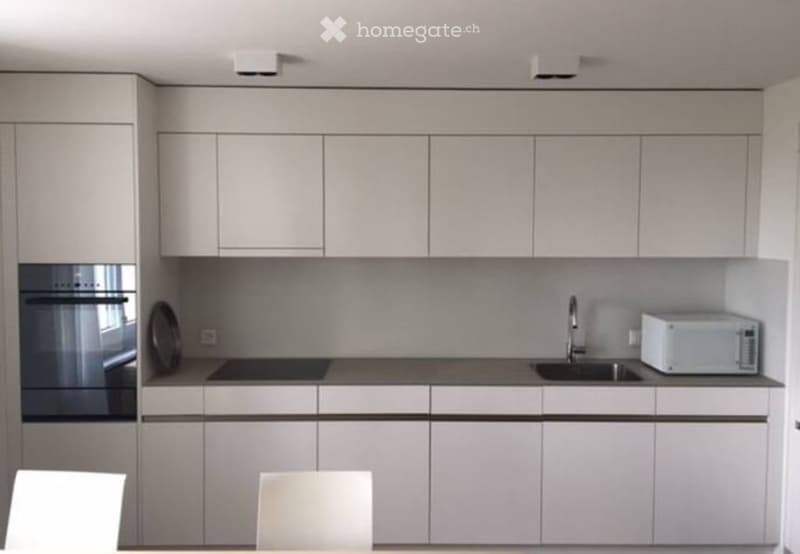 Modern kitchen with white cabinets, stainless steel sink, microwave, and two white chairs at the counter.