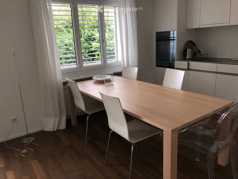 Dining area with a light wooden table, white and transparent chairs, and a modern oven in the background.