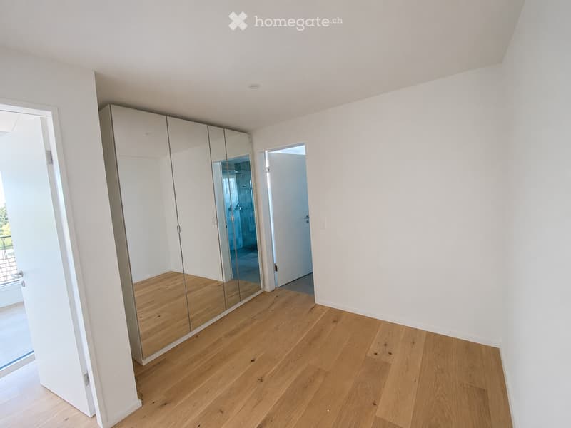 Empty room with light wooden flooring, white walls, and a large mirrored wardrobe. A door leads to a bathroom with blue tiles visible.