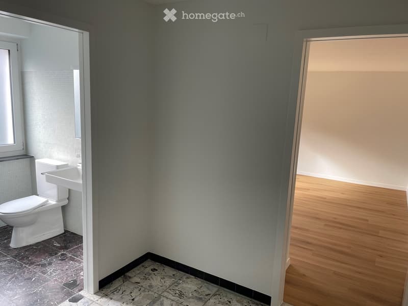 Bathroom with a toilet, sink, and tiled floor. The room has a grey wall and a wooden floor in the adjacent area.